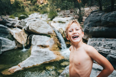Portrait of happy shirtless boy standing by stream in forest