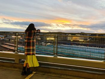 Rear view of woman standing by railing against sky during sunset