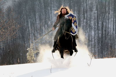 Full length of man with sword riding horse on snow covered field