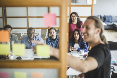 Smiling colleagues looking at businessman explaining strategy in meeting seen through glass wall