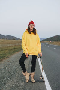 Full length of woman holding skateboard standing on road against sky