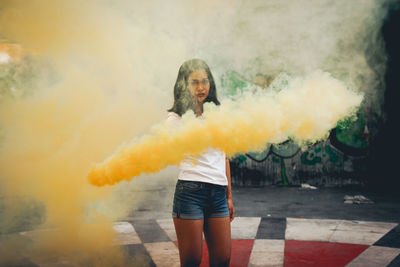 Portrait of woman holding yellow distress flare while standing on footpath