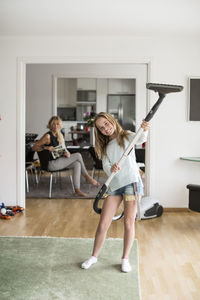 Full length portrait of playful girl holding vacuum cleaner at home with mother sitting in background