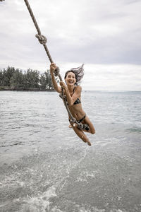 Happy woman swinging on rope over sea against cloudy sky