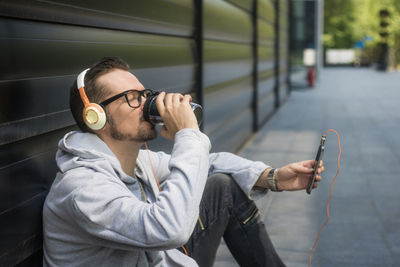 Man listening music on mobile phone while drinking on footpath