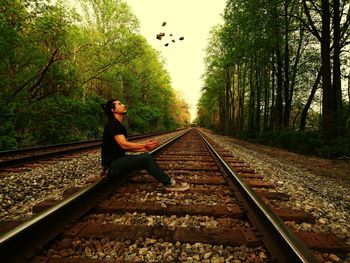 Man sitting on railroad track looking at stone in mid-air against trees