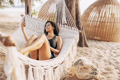 Smiling woman doing video on smart phone while lying down in hammock at beach
