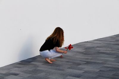 Full length of woman with flowers crouching on footpath by white wall