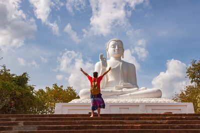 Rear view of man with arms raised standing by giant buddha statue