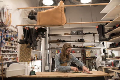 Shoemaker cutting leather on workbench surrounded by shoes at workshop