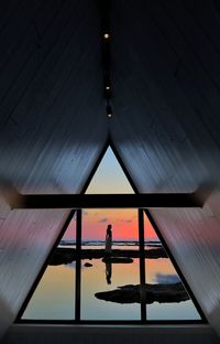 Silhouette woman standing at beach seen through window during sunset