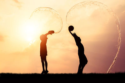 Silhouette friends splashing water with containers while standing on field against orange sky