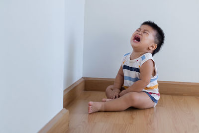Boy crying while sitting on hardwood floor against wall