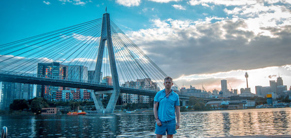 Portrait of man standing by river against suspension bridge