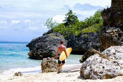 Rear view full length of shirtless man with surfboard walking at beach on sunny day