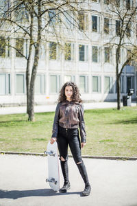 Full length portrait of confident teenage girl standing with skateboard on street against building