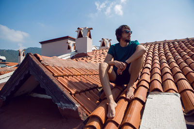 Man in the green t-shirt shorts and sunglasses sitting on the roof of a clay orange tile