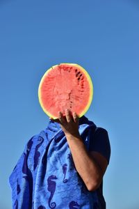 Man covering face with watermelon while standing against clear sky