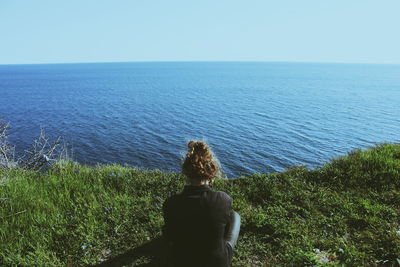 Woman sitting on grass cliff overlooking calm sea against clear sky