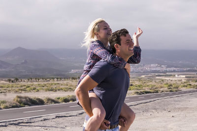 Side view of man piggybacking girlfriend on road against sky