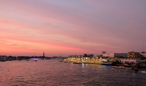 Illuminated buildings by river against sky during sunset