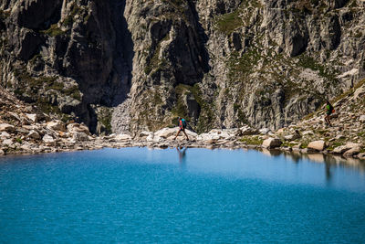 Distant unrecognizable adventurer with backpack walking alone on rough stony shore between blue lake and steep rock in pyrenees