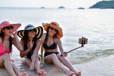 Female friends in hat and sunglasses taking selfie at beach