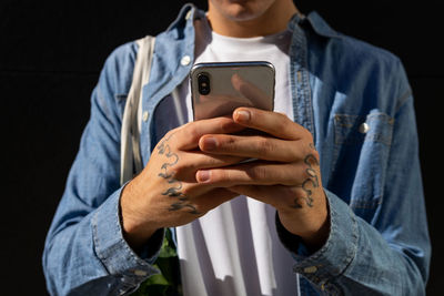 Cropped unrecognizable modern young male browsing mobile phone while standing against black wall on street