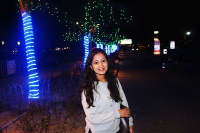 Portrait of smiling young woman standing against illuminated trees at night