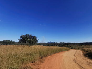 Dirt road amidst field against clear blue sky