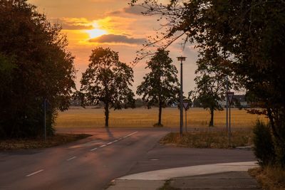 Road by trees against sky during sunset