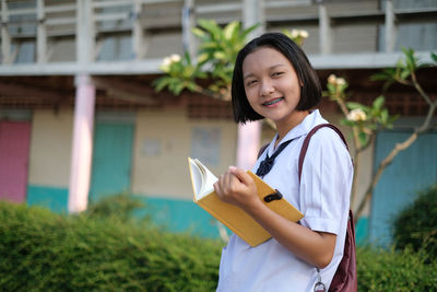 Smiling teenage girl holding book while standing outdoors