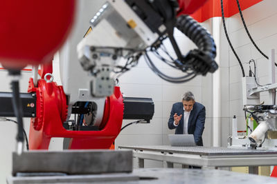 Mature male entrepreneur discussing over laptop during video conference while machinery in foreground