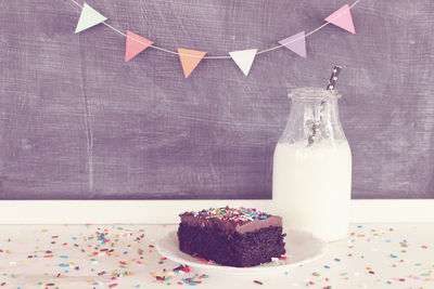 Lit candle on birthday cake with by milk bottle against decorated wall