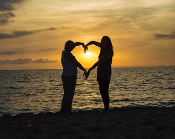 Silhouette women making heart shape while standing on shore during sunset
