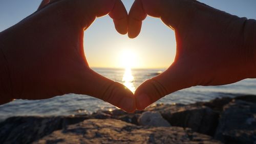 Cropped image of hand making heart shape against sea during sunset