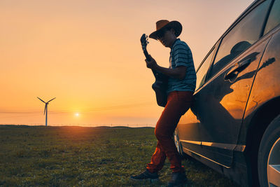 Mature man playing guitar by car on field during sunset