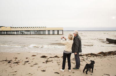 Full length of senior woman photographing while standing by man holding dog's leash at beach against clear sky