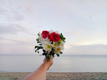 Cropped hand of person holding flower bouquet against sea and sky