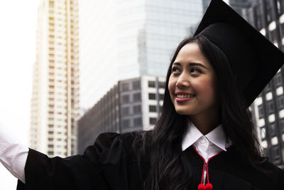 Smiling young woman wearing graduation gown in city