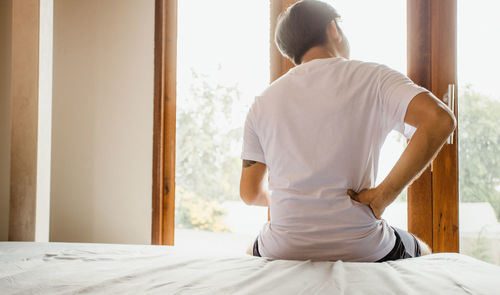 Rear view of man sitting on bed at home