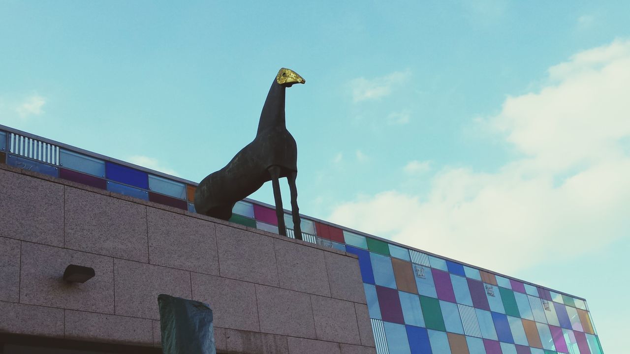 low angle view, sky, bird, animal themes, blue, one animal, built structure, perching, architecture, art and craft, animals in the wild, wildlife, sculpture, animal representation, art, day, outdoors, cloud - sky, building exterior, human representation