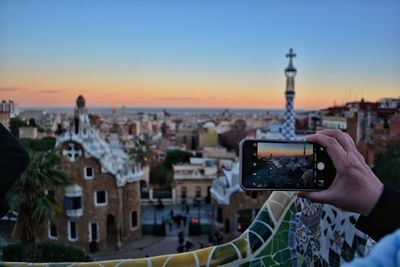 Cropped image of hand photographing church with cityscape during sunset