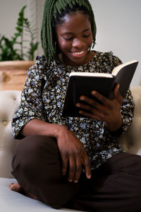 Positive african american female with braids reading interesting book while resting on couch in modern living room at home