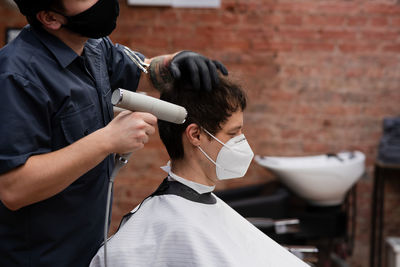 Barber in protective mask and latex glove styling adult man in a barbershop salon. new normal