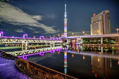 Illuminated bridge over river by buildings against sky at night