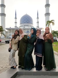 Portrait of smiling friends showing thumbs up while standing against mosque