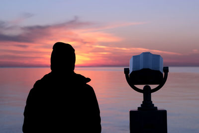 Rear view of person standing by coin-operated binoculars looking at sea during sunset