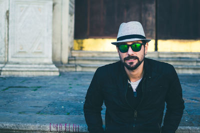 Portrait of young man wearing hat while standing against built structure