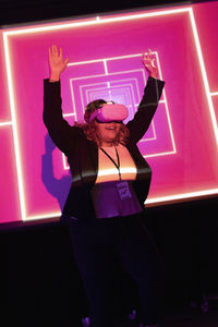 Young businesswoman with arms raised watching through vr glasses at illuminated workshop